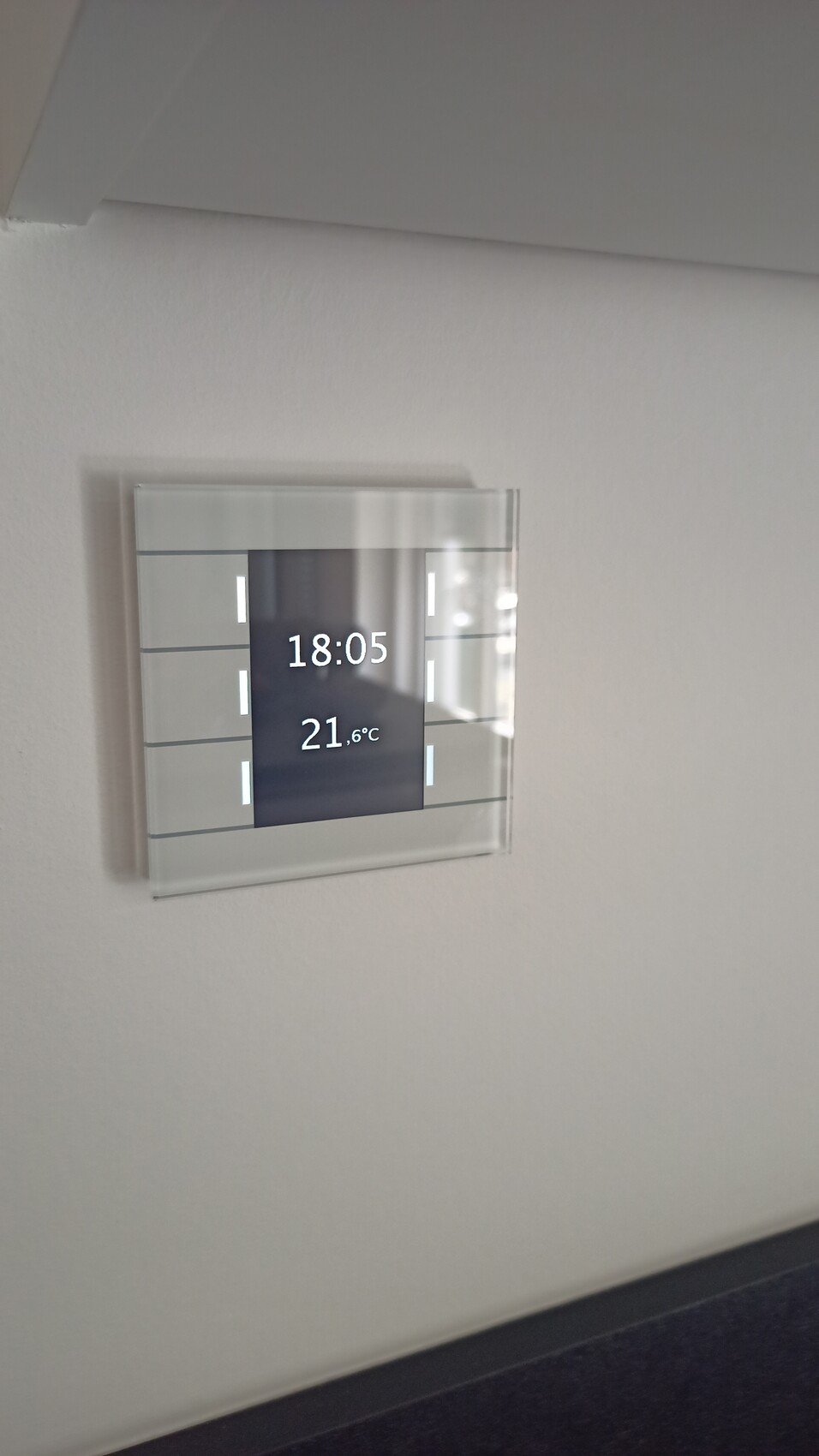 Digitales Wanddisplay zeigt Uhrzeit 18:05 und Temperatur 21,6°C in einem Glasrahmen.