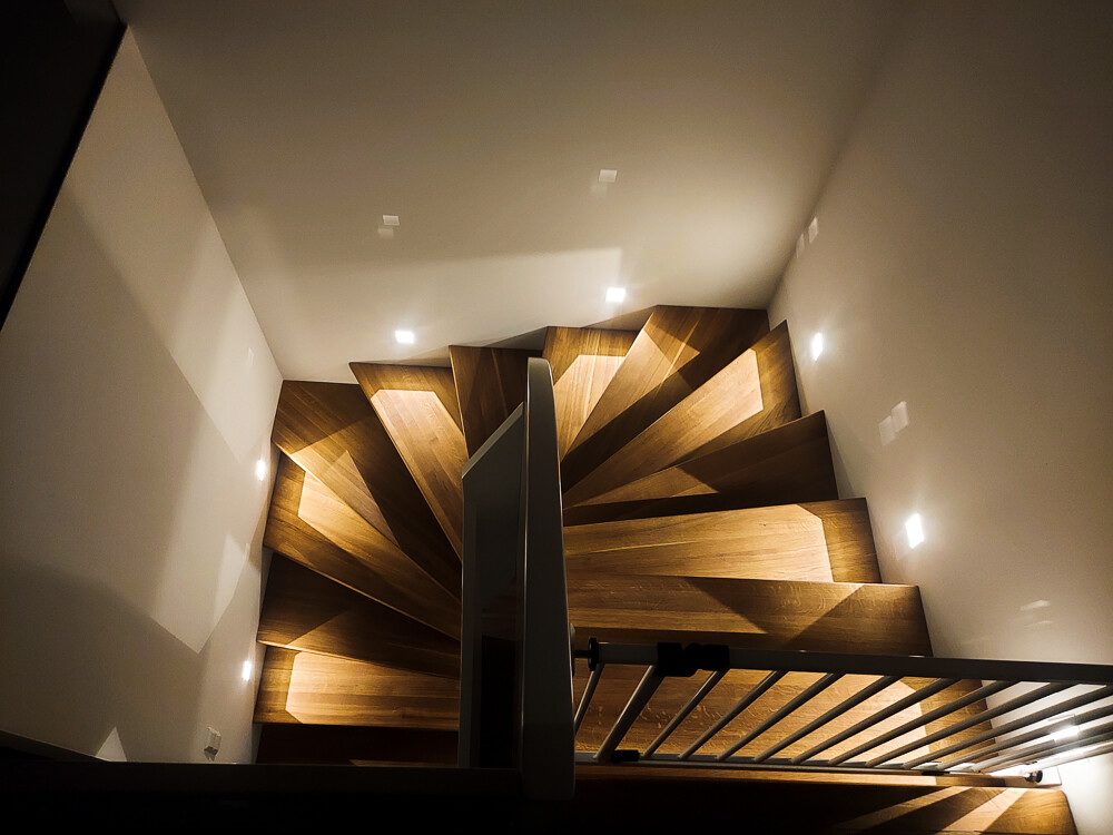 Spiralholztreppe von oben, warm beleuchtet, weiße Wände, Geländer am Rand.