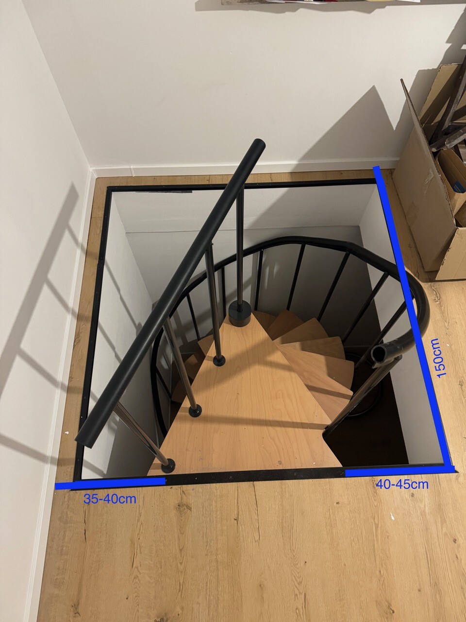 Spiralholztreppe in offenem Loch; Holzstufen, schwarzes Geländer, Kartonbox daneben.