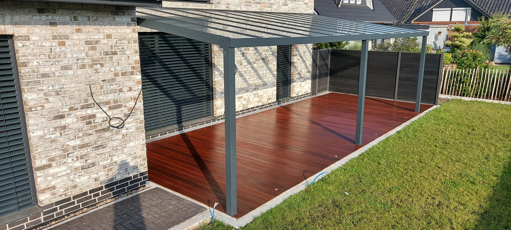 Überdachte Holzterrasse am Haus mit rotem Boden, grauem Metallrahmen und dunklen Sichtschutzwänden.