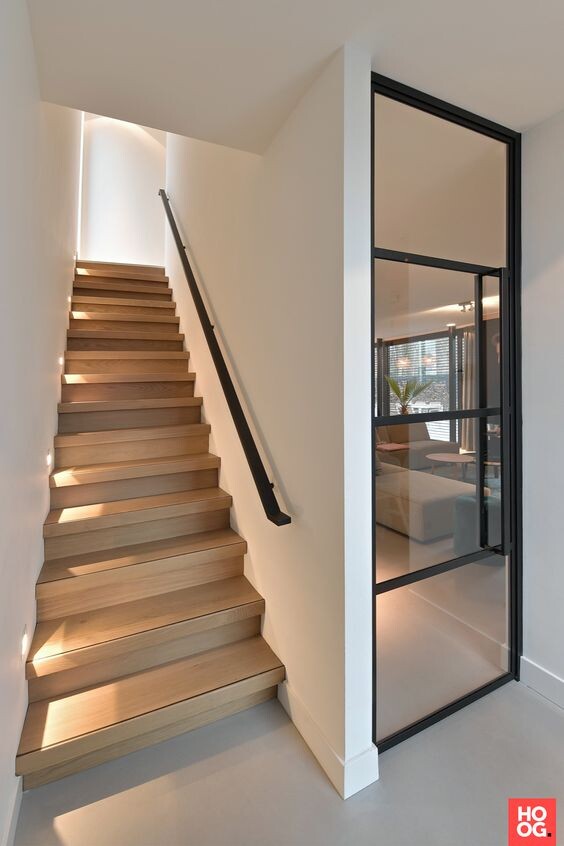 Moderne Holztreppe links mit schwarzem Geländer, Glastür rechts, Blick ins Wohnzimmer.