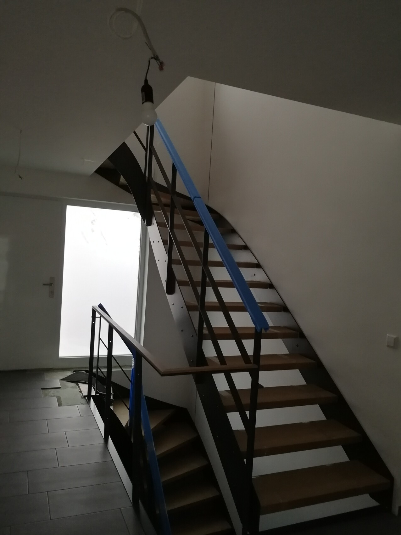 Gebogene Metalltreppe mit Holzstufen, blaue Folie am Geländer, Baustellenbeleuchtung.