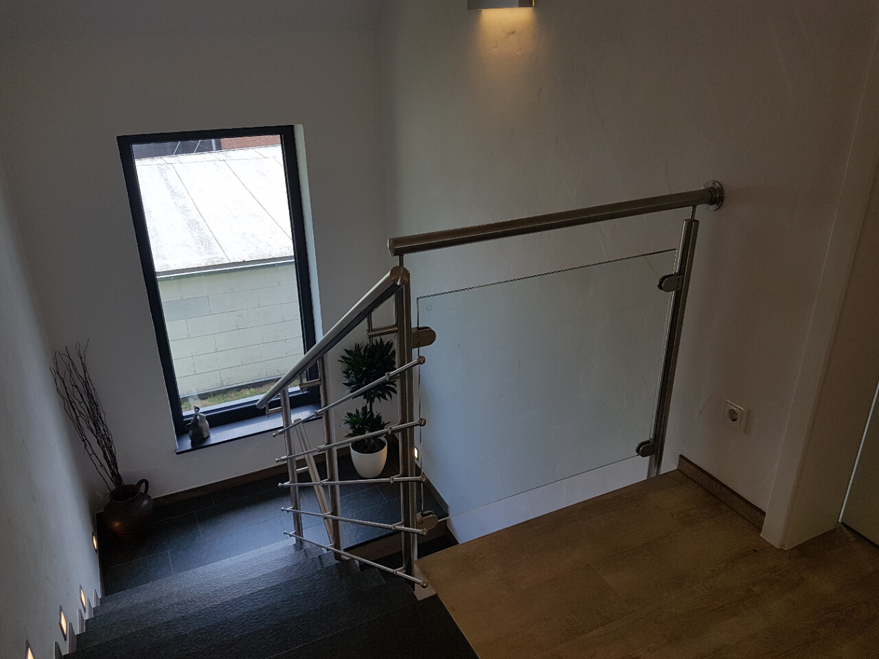 Innenansicht einer Treppe mit Glasgeländer, dunklen Stufen, Fenster links und Pflanze im Topf