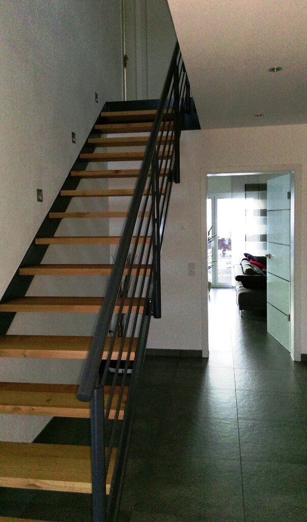 Moderne Innenansicht mit Holzstufen-Treppe und Metallgeländer links; Flur führt zum hellen Wohnzimmer.