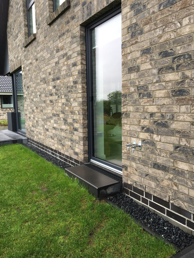 Außenansicht eines Backstein-Gebäudes mit großem Fenster, Metallstufe und Wasserhahn an der Wand