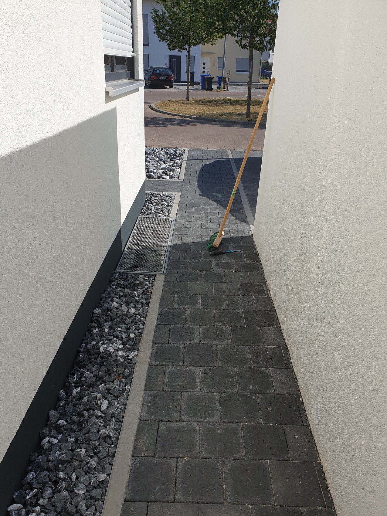 Enge, gepflasterte Gasse zwischen weißen Hauswänden; Besen lehnt an Wand, Auto im Hintergrund.