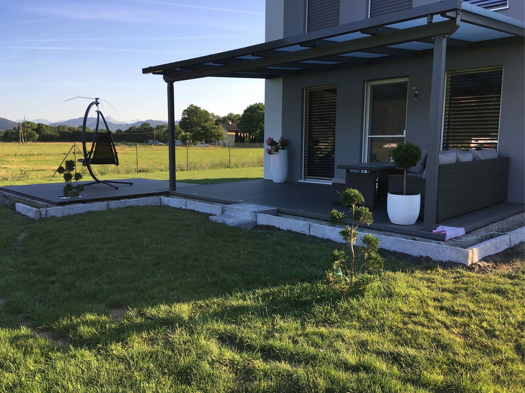 Überdachte Terrasse am modernen Haus mit Lounge-Sofa, Pflanzkübeln und Schaukelstuhl.