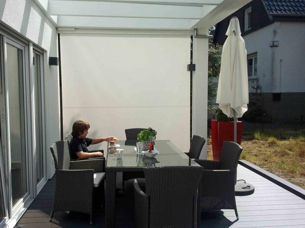 Gedeckter Terrassentisch aus Glas mit Stuehlen, Sonnenschirm und Garten hinter dem Haus.