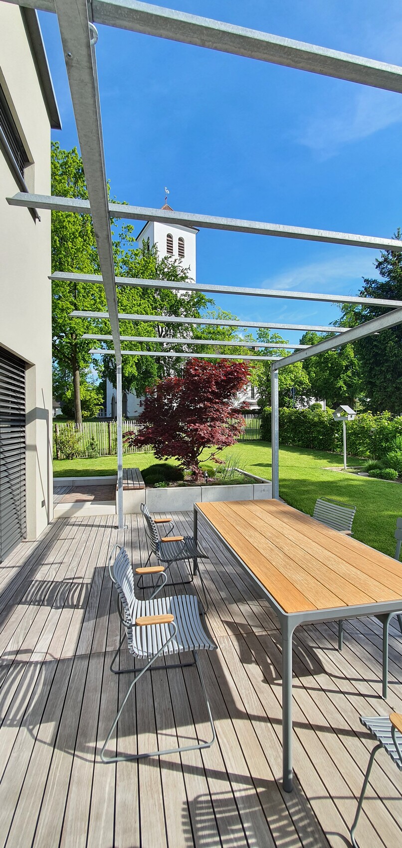 Garten-Terrasse mit Holztisch, Metallstühlen, grünem Garten und Kirchturm im Hintergrund