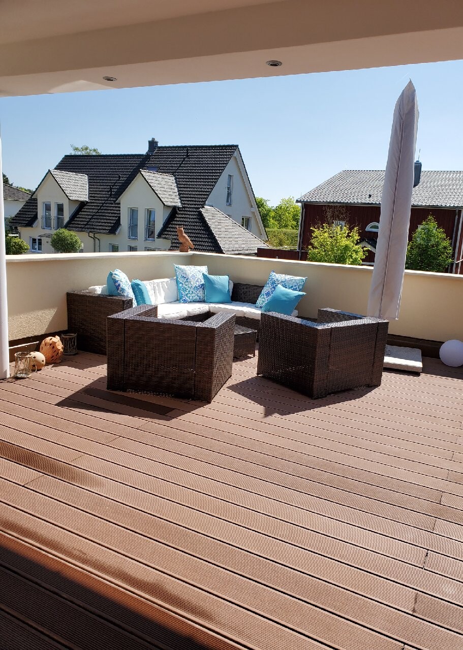 Terrasse mit dunkler Rattan-Eckcouch, blauen Kissen, Glas-Tisch und Sonnenschirm; Nachbarhäuser.