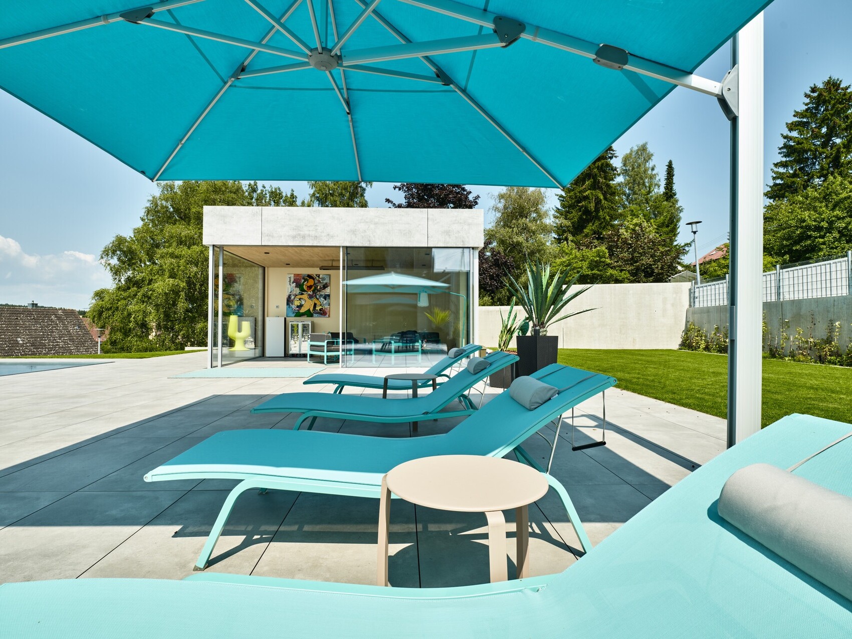 Poolbereich mit türkisfarbenen Liegen, großem Sonnenschirm und modernem Glasgebäude im Hintergrund.