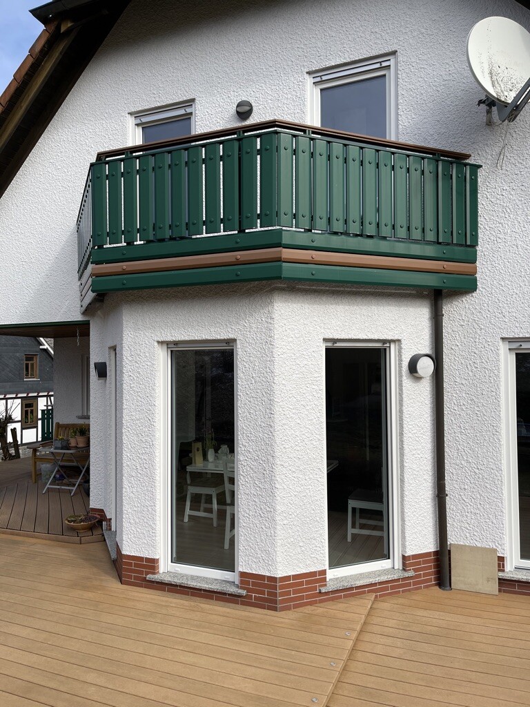 Zweigeschossiges Haus mit grünem Balkongeländer, Glasfronten und Holzterrasse.