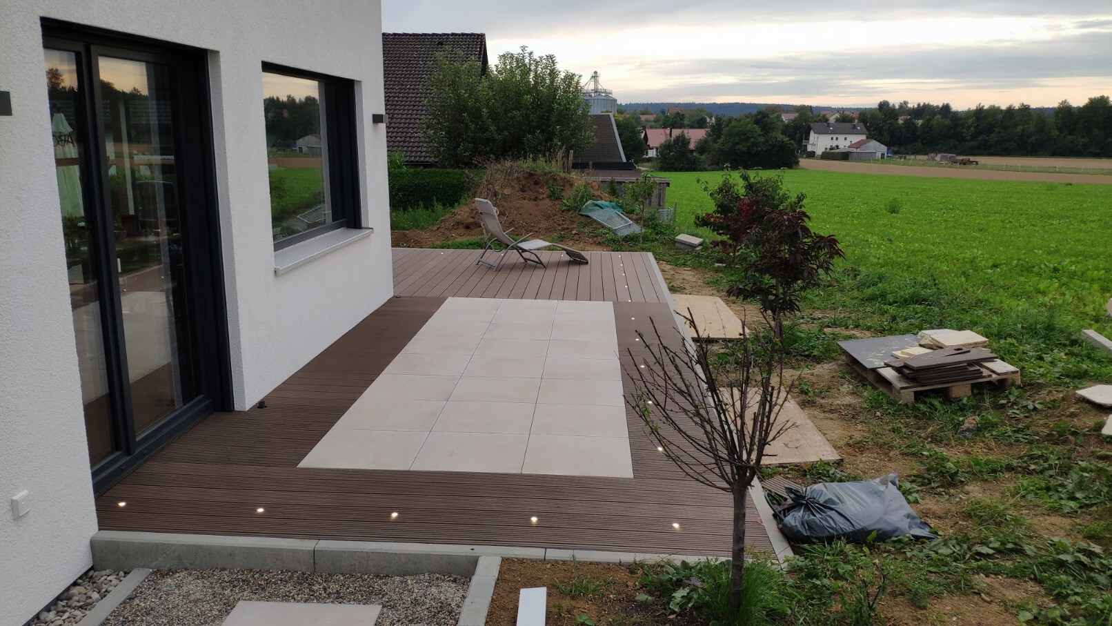 Moderne Holzterrasse mit hellen Fliesen und Lounge-Stuhl, Blick auf grüne Felder.