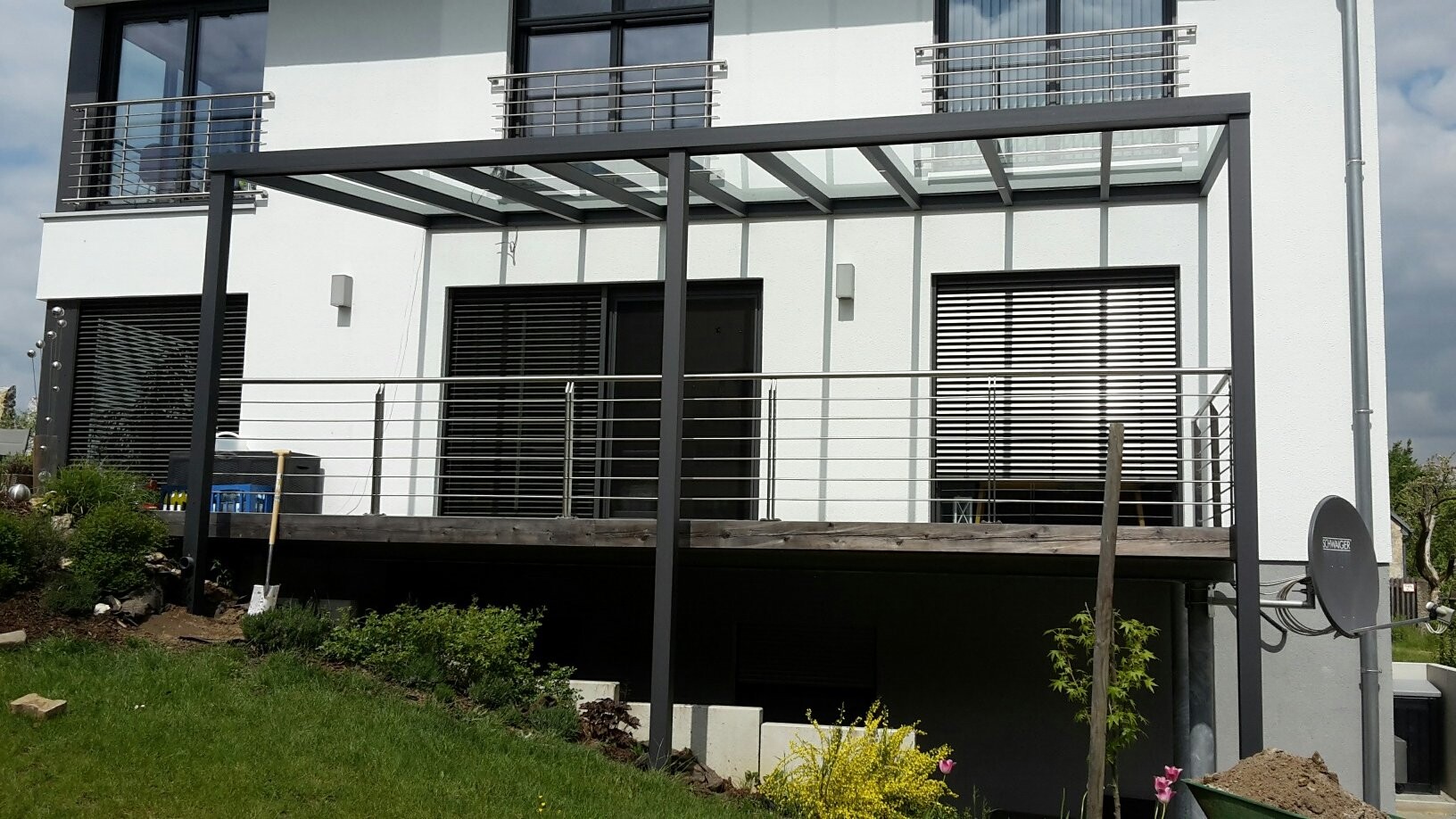 Moderne weiße Hausfassade mit Glas-Pergola über Terrasse und Geländer; Garten im Vordergrund.