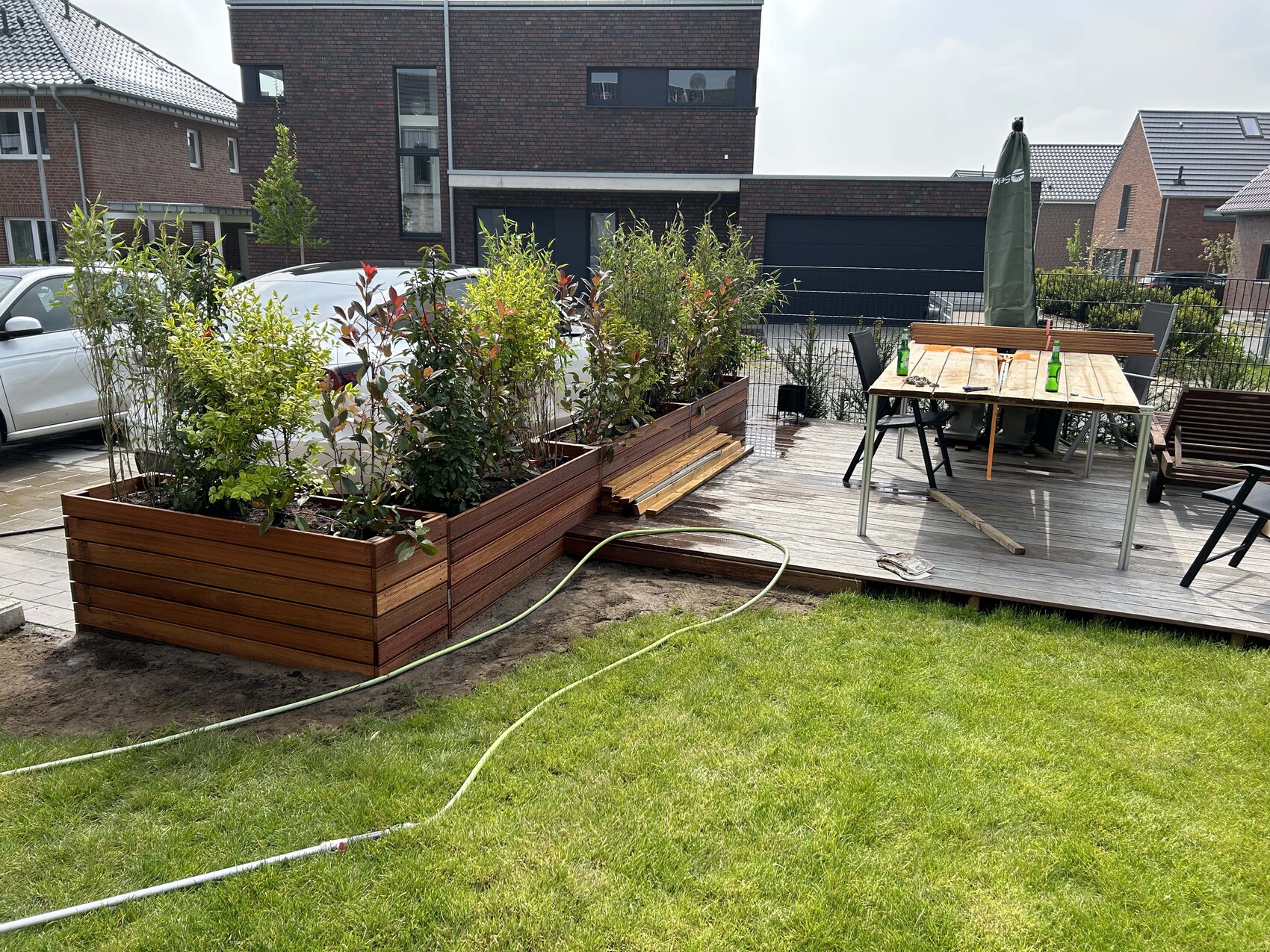 Holzdeck mit Pflanzkästen, Esstisch mit Stühlen, grünem Schirm und Gartenschlauch.