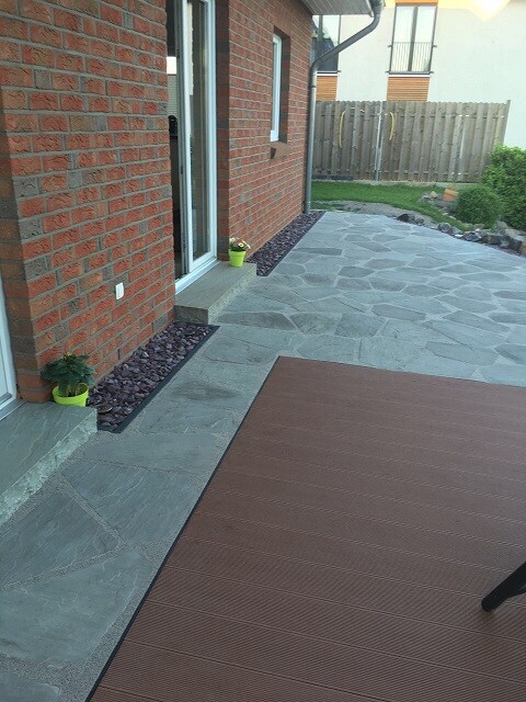 Garten-Terrasse mit grauen Pflastersteinen, Holzdeck und gelben Pflanzkübeln neben dem Haus.