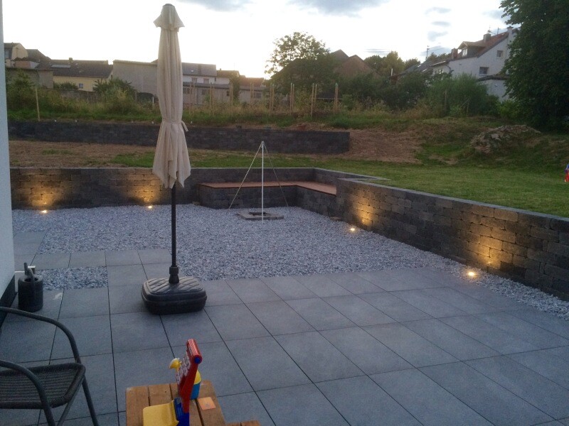Garten-Terrasse mit grauen Fliesen, geschlossener Sonnenschirm, Spielzeug und beleuchteter Steinmauer.