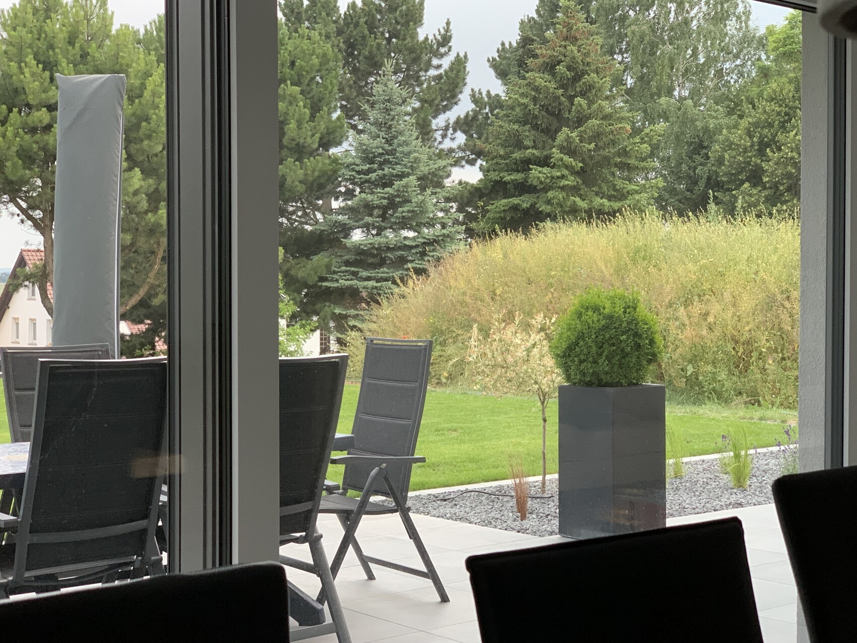 Blick von innen auf Terrasse: schwarze Stühle um einen Tisch, Glasfront, grüner Garten.
