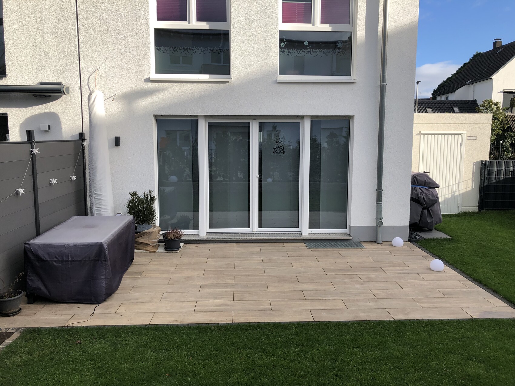 Gartenterrasse vor weißem Haus mit Glasschiebetüren, Holzdielen und beleuchteten Kugeln.