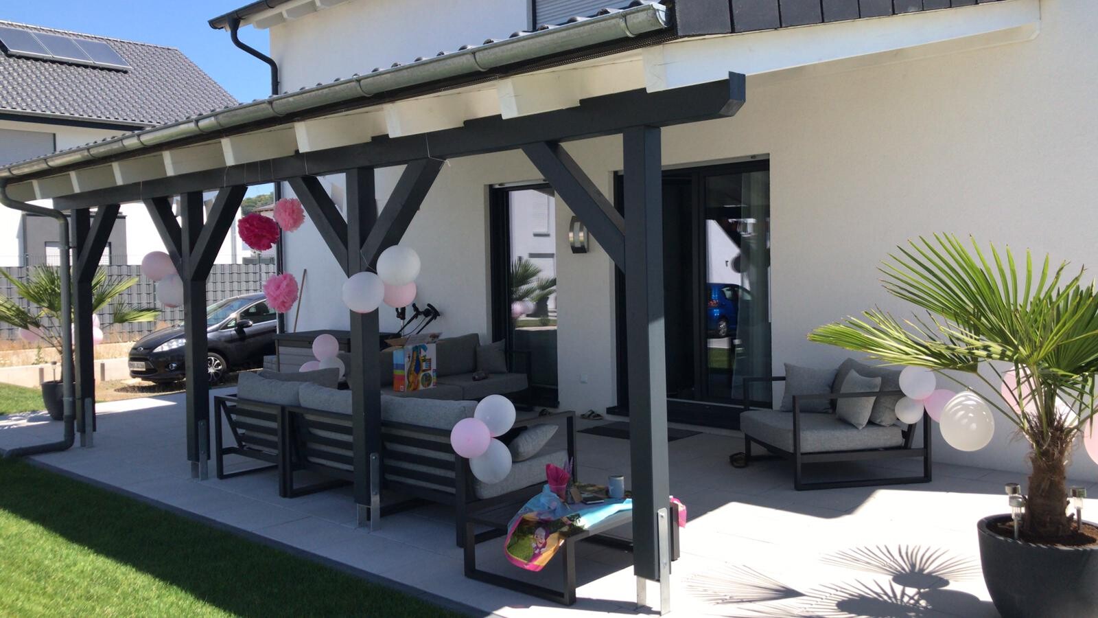 Überdachter Patio mit dunklem Holzgerüst, rosa-weiße Ballons und Gartenmobiliar.