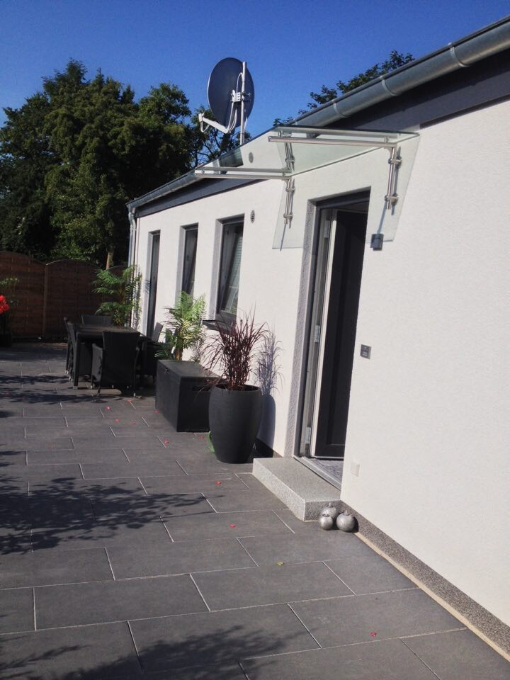 Weiße Hausfassade mit Glasvordach über Tür, Satellitenschüssel, Pflanzkübel und Terrassenfliesen