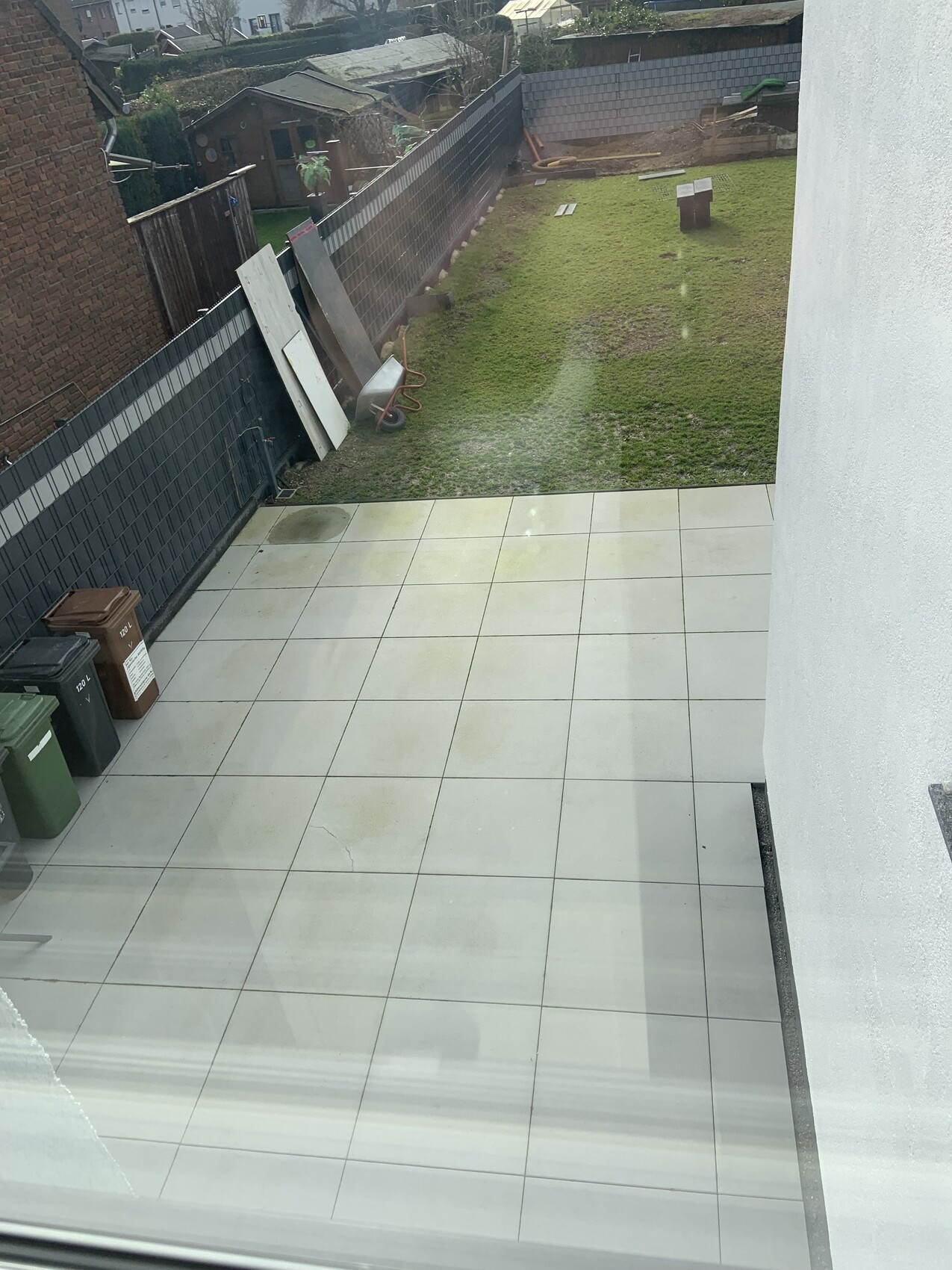 Blick von oben auf helle Fliesen-Terrasse; links Müllbehälter, dahinter Garten mit Hütte.