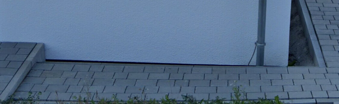 Außenansicht: hellblaue Wand, graue Pflastersteine, Abflussrohr, Randbegrünung.