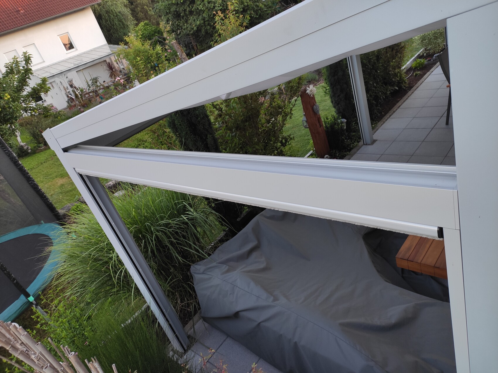 Weiße Metallkonstruktion einer überdachten Terrasse mit grauer Abdeckung, Garten im Hintergrund.