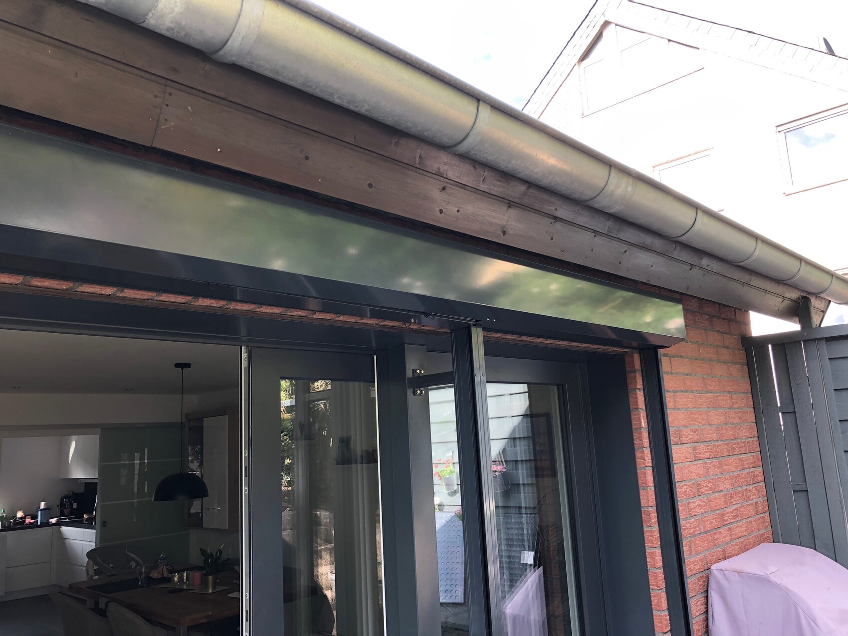Offene Glas-Schiebetüren führen ins Esszimmer; außen Backsteinwand, Dachrinne und Holzbalken.