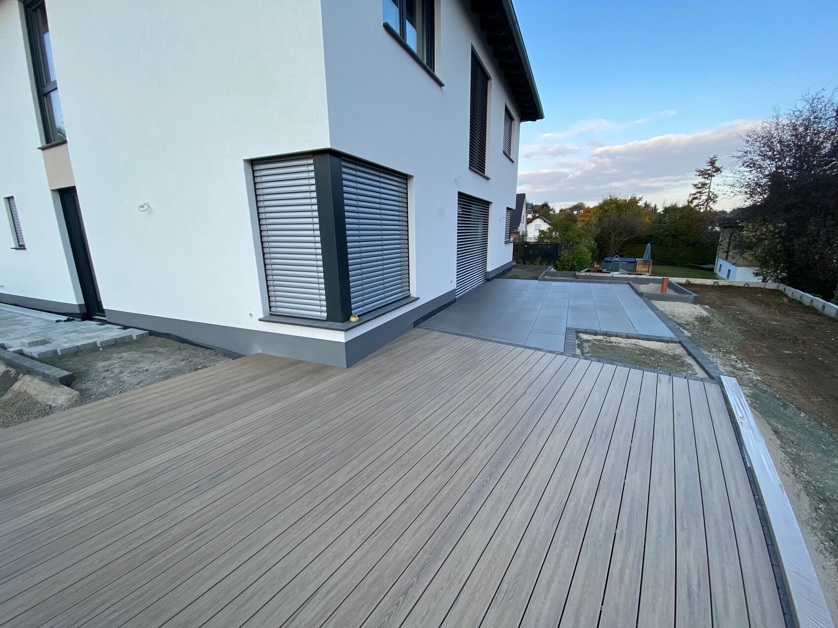 Moderne weiße Hausfassade mit Rollläden, große Holzterrasse im Vordergrund und Garten im Hintergrund