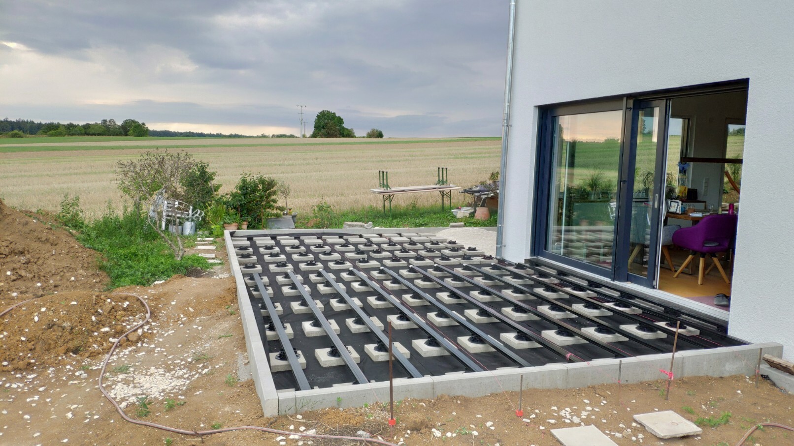 Außenansicht eines Hauses mit Glasfront; Terrassenunterbau im Bau vor Feldern.