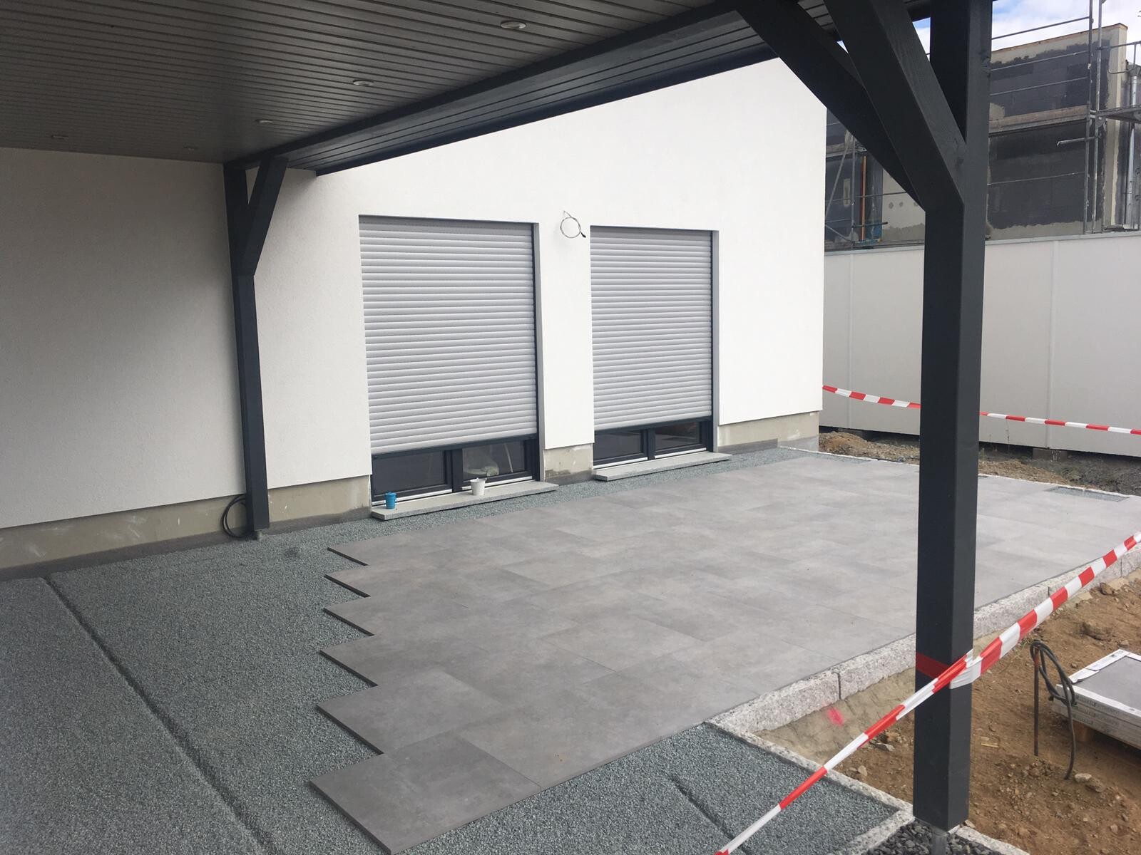 Moderne Terrasse neben Neubau: weiße Wand mit zwei Rollläden, graue Bodenplatten, Absperrband.