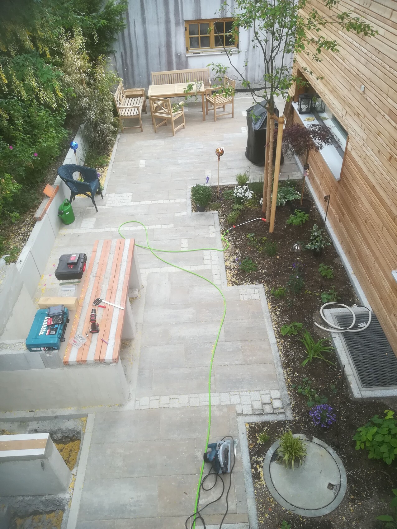 Garten-Terrasse mit Holzmöbeln, Bauwerkzeugen, grünem Schlauch und Pflanzenbeete.