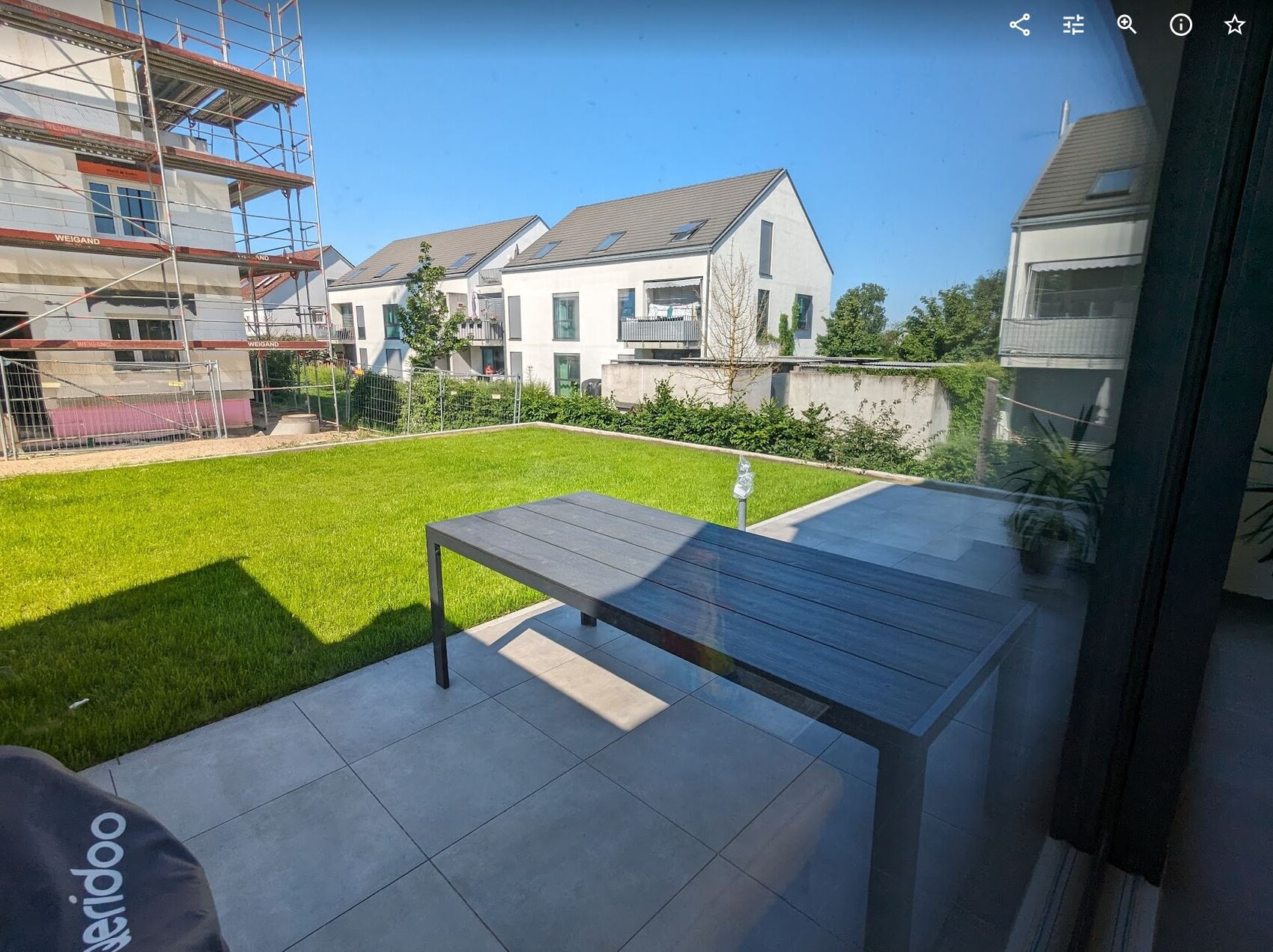 Blick durch Glasfront auf Terrasse mit dunklem Tisch, grünem Rasen und modernen Häusern; Baugerüst links.