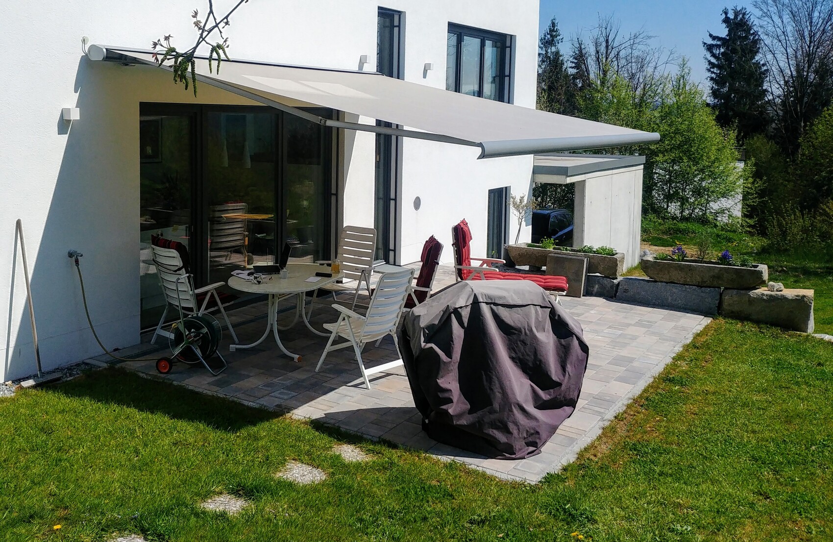 Weiße Hausfassade mit Terrasse, Markise, runder Tisch, Stühle und abgedeckter Grill.