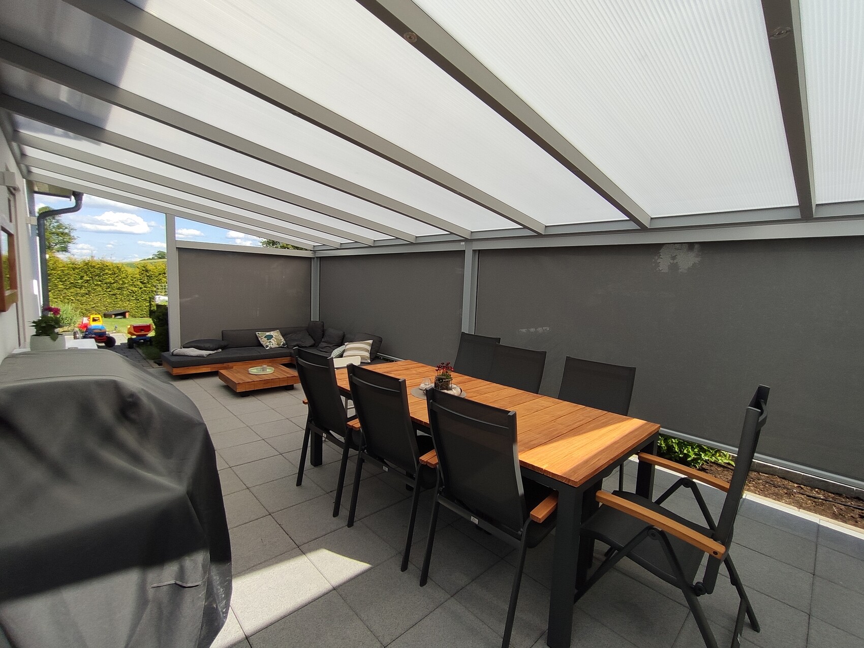 Überdachte Terrasse: langer Holztisch, schwarze Stühle, Lounge-Ecke, Gartenblick