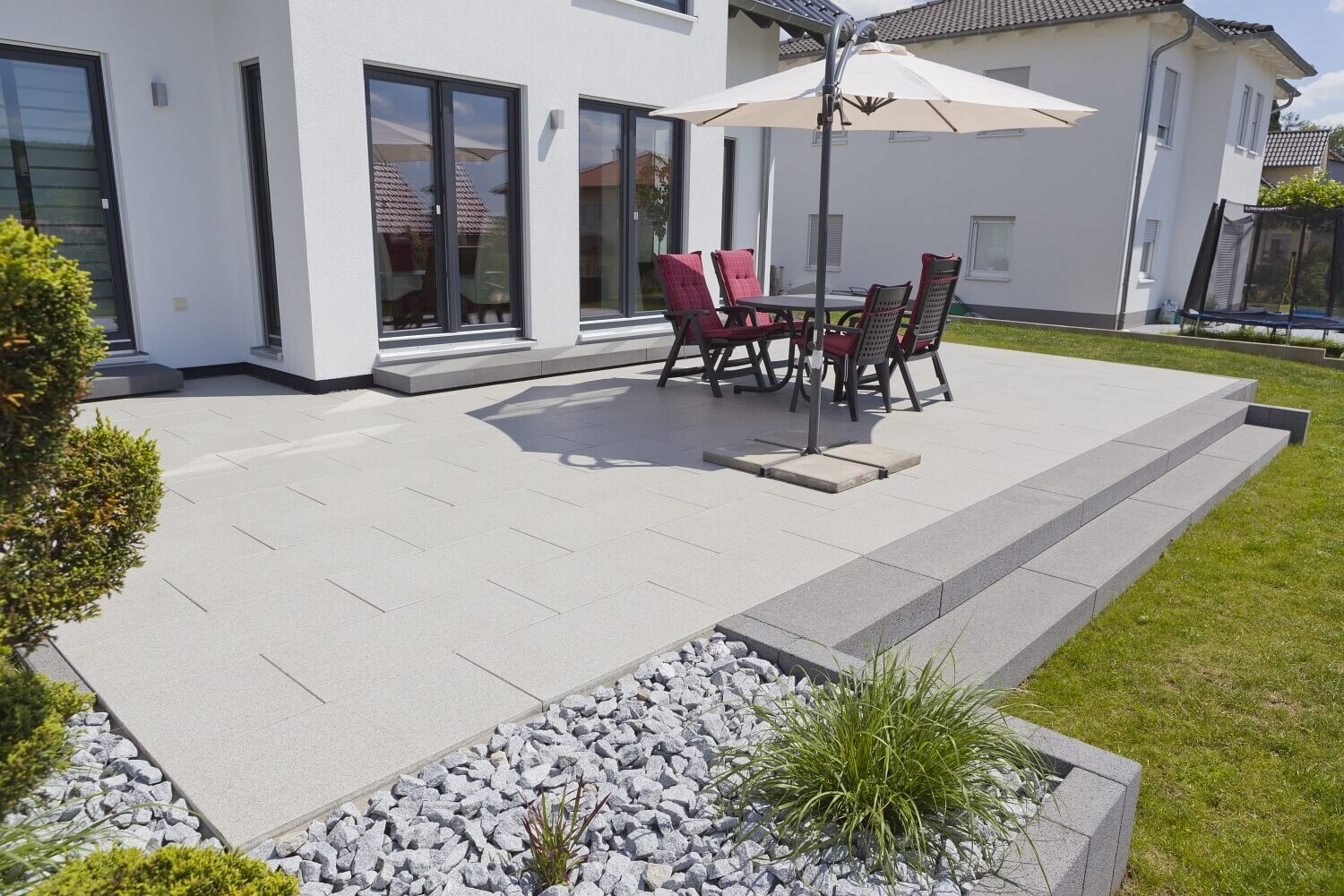 Moderne Terrasse am weißen Haus mit hellgrauen Pflasterplatten, Esstisch, Stühle und Sonnenschirm.