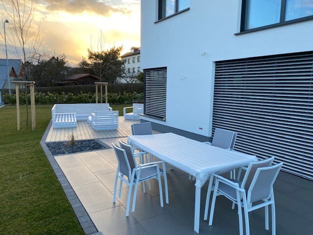 Moderne Terrasse am weißen Haus mit Esstisch und Stühlen, Lounge-Bereich und Sonnenuntergang.