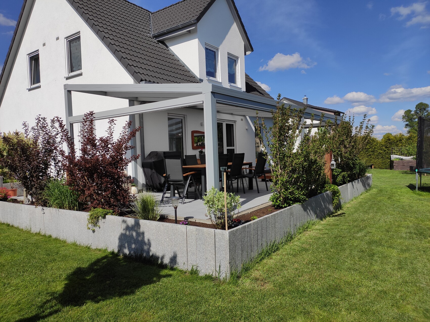 Weißes Haus mit Pergola-Terrasse, Esstisch und Stühle im Garten.