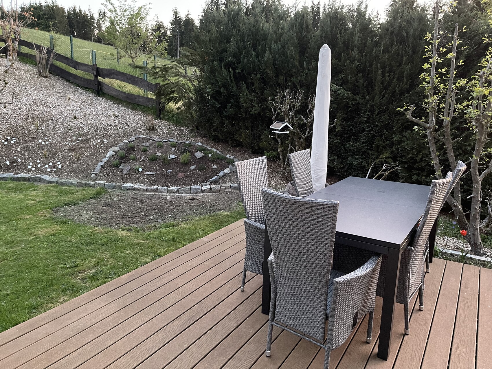 Holzdeck mit grauem Gartentisch und wicker-Stühlen, geschlossener weißer Sonnenschirm, Hecke dahinter.
