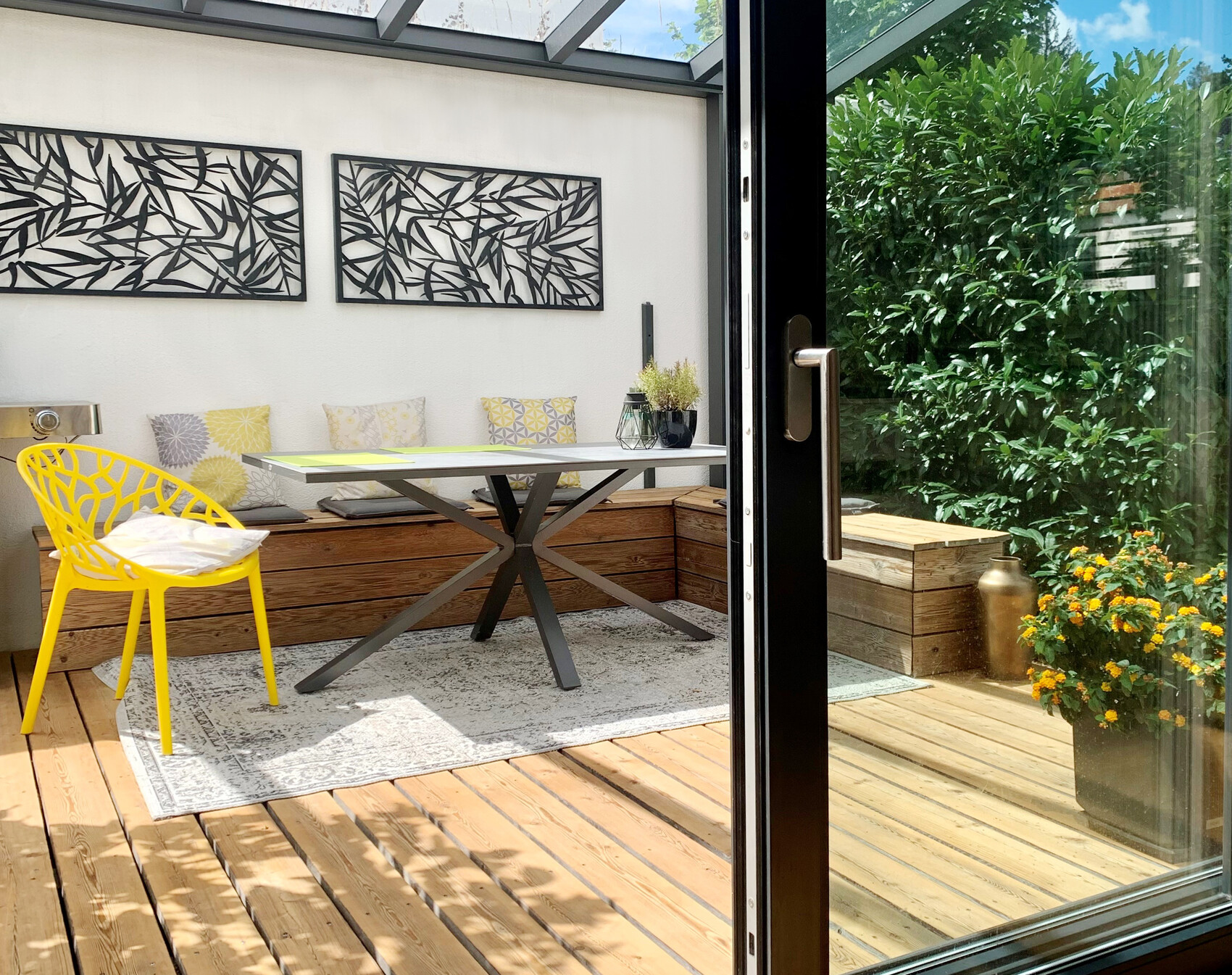 Garten-Patio mit gelbem Stuhl, Glastisch, gepolsterter Bank, Wandkunst, Glas-Schiebetür.