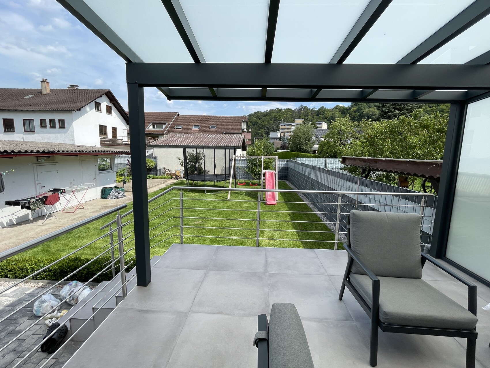 Moderne Balkonterrasse mit grauen Loungesessel, Glasdach, Blick auf Garten mit Spielgerät wie Rutsche.