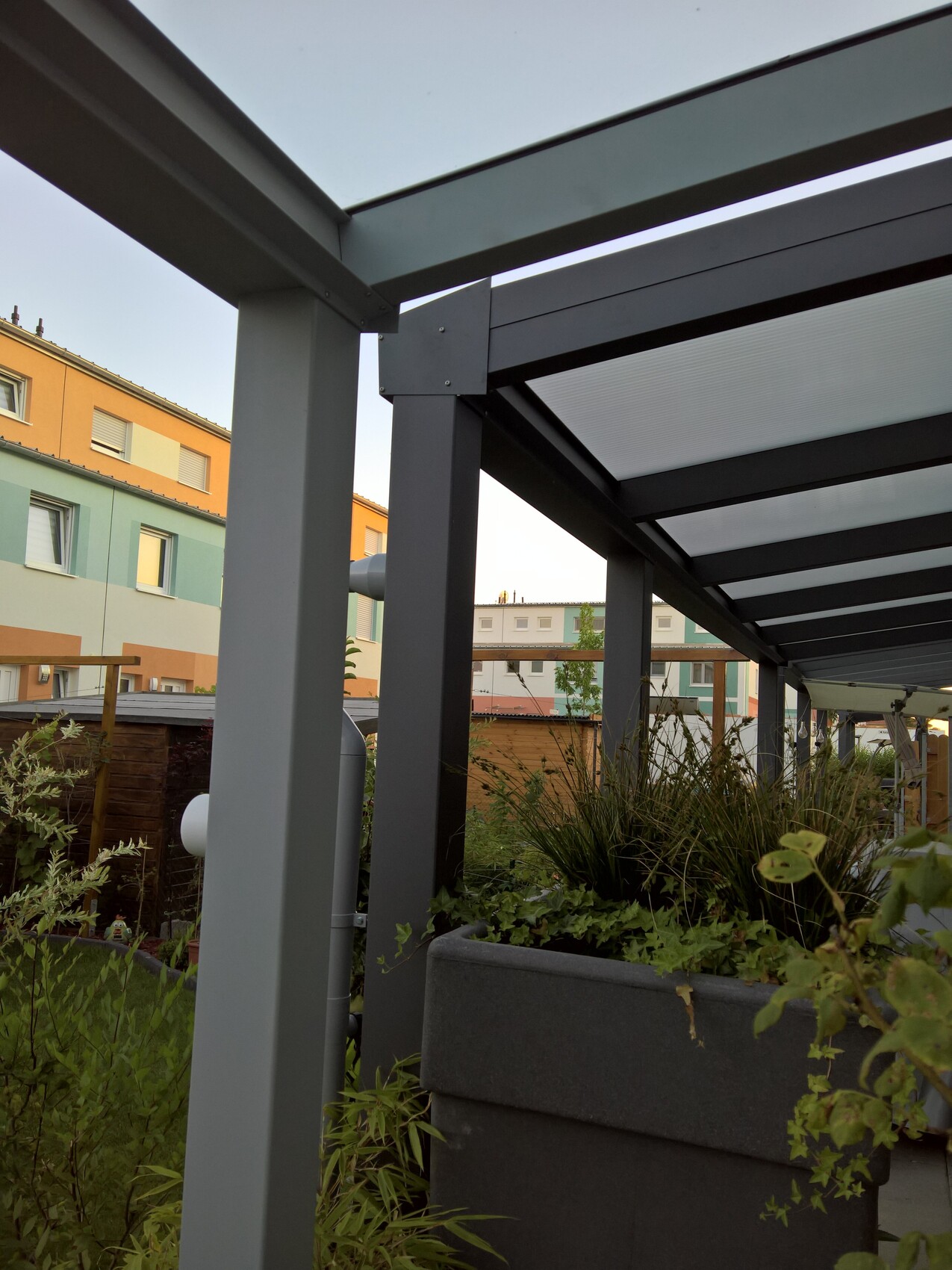 Pergola aus Metallrahmen mit Glasdach über Pflanzkübel, farbige Häuser im Hintergrund.