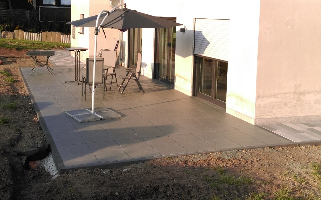Gepflasterte Terrasse am Haus mit Sonnenschirm, Tisch und Stühlen.
