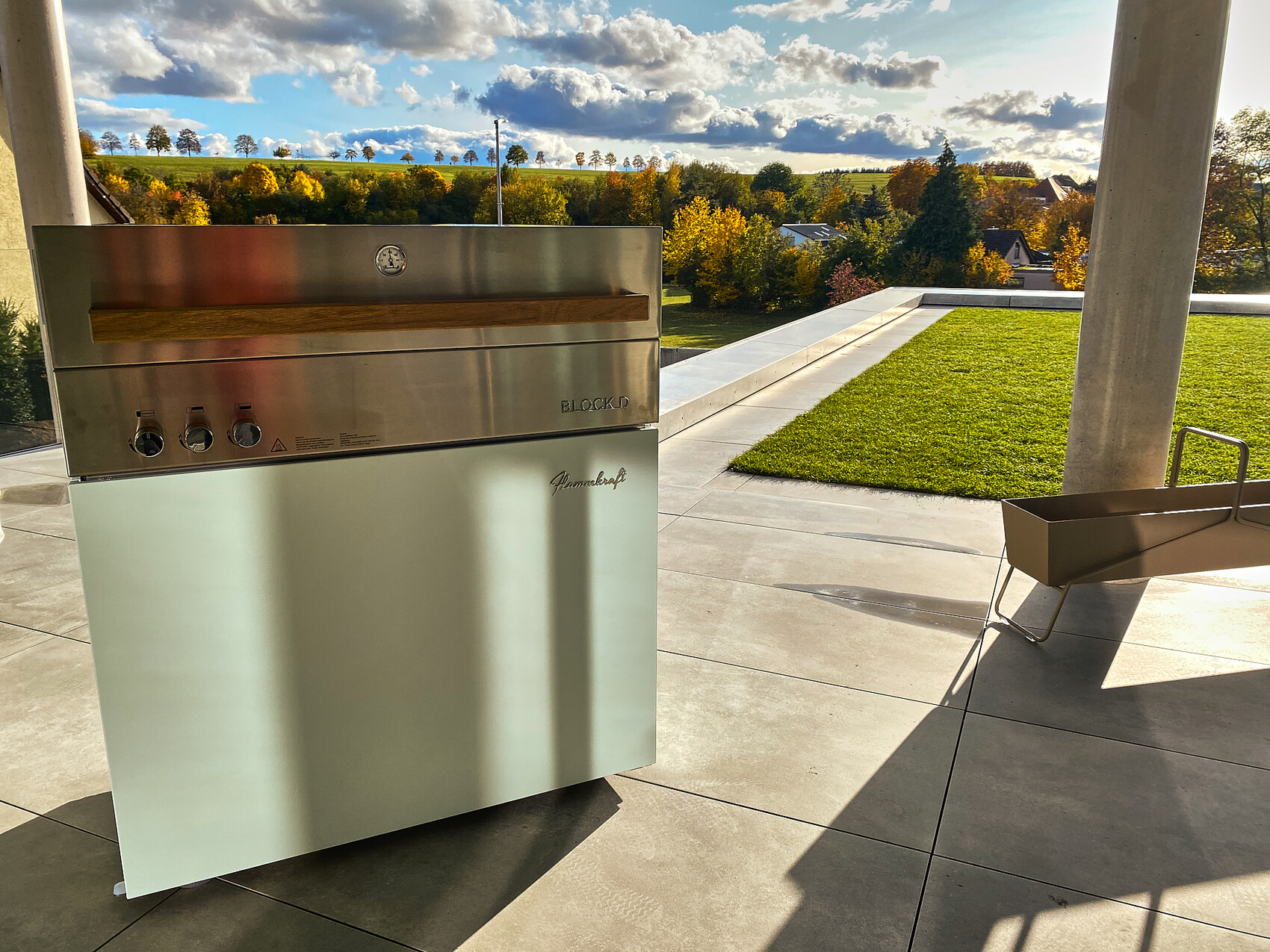 Außen-Edelstahl-Gasgrill mit Holzgriff auf Terrasse; Herbstlandschaft mit bunten Bäumen.