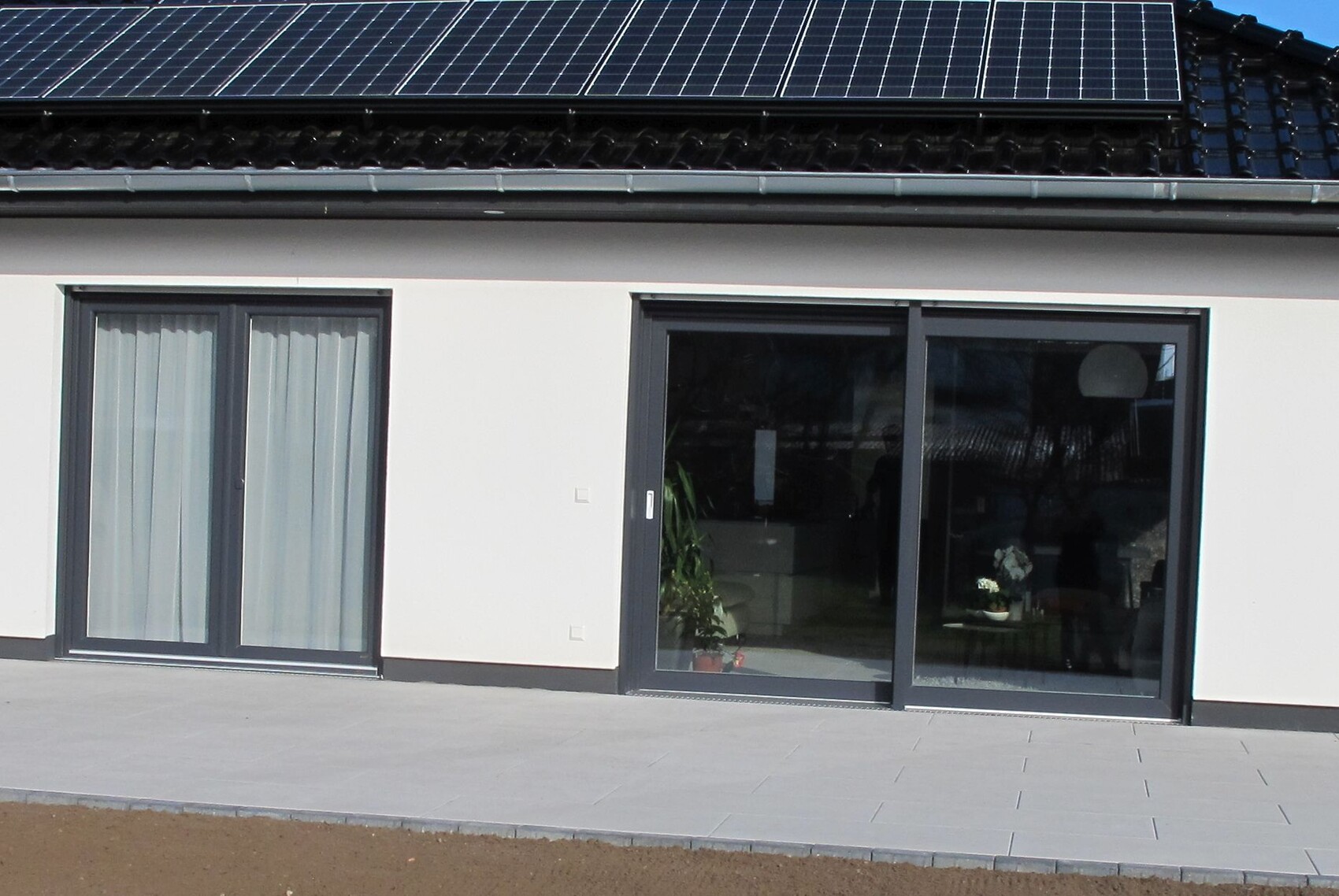 Moderne Hausfassade mit Solarpanelen auf dem Dach und großen Glas-Schiebetüren zur Terrasse.