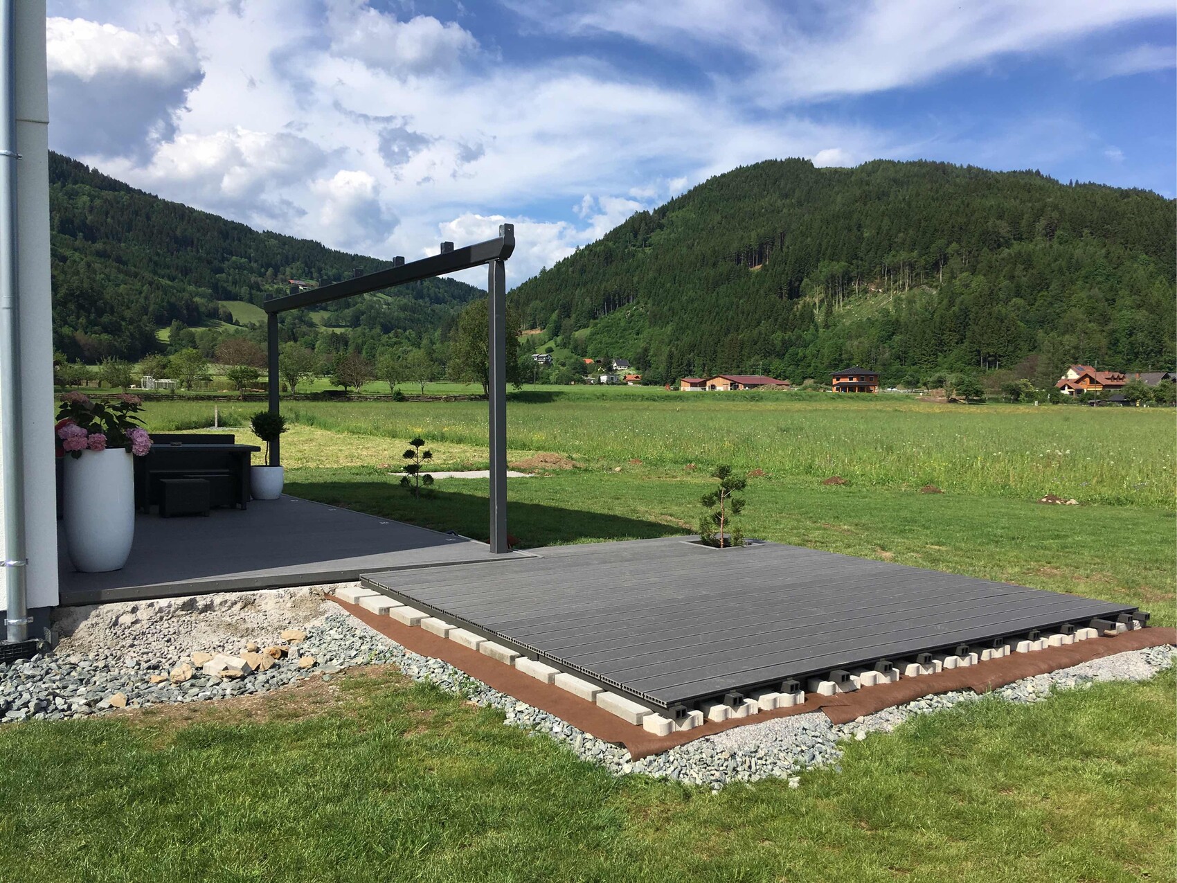 Terrasse mit dunkler Holzplatte und Pergola, Topfpflanzen, grüne Felder und Berglandschaft.
