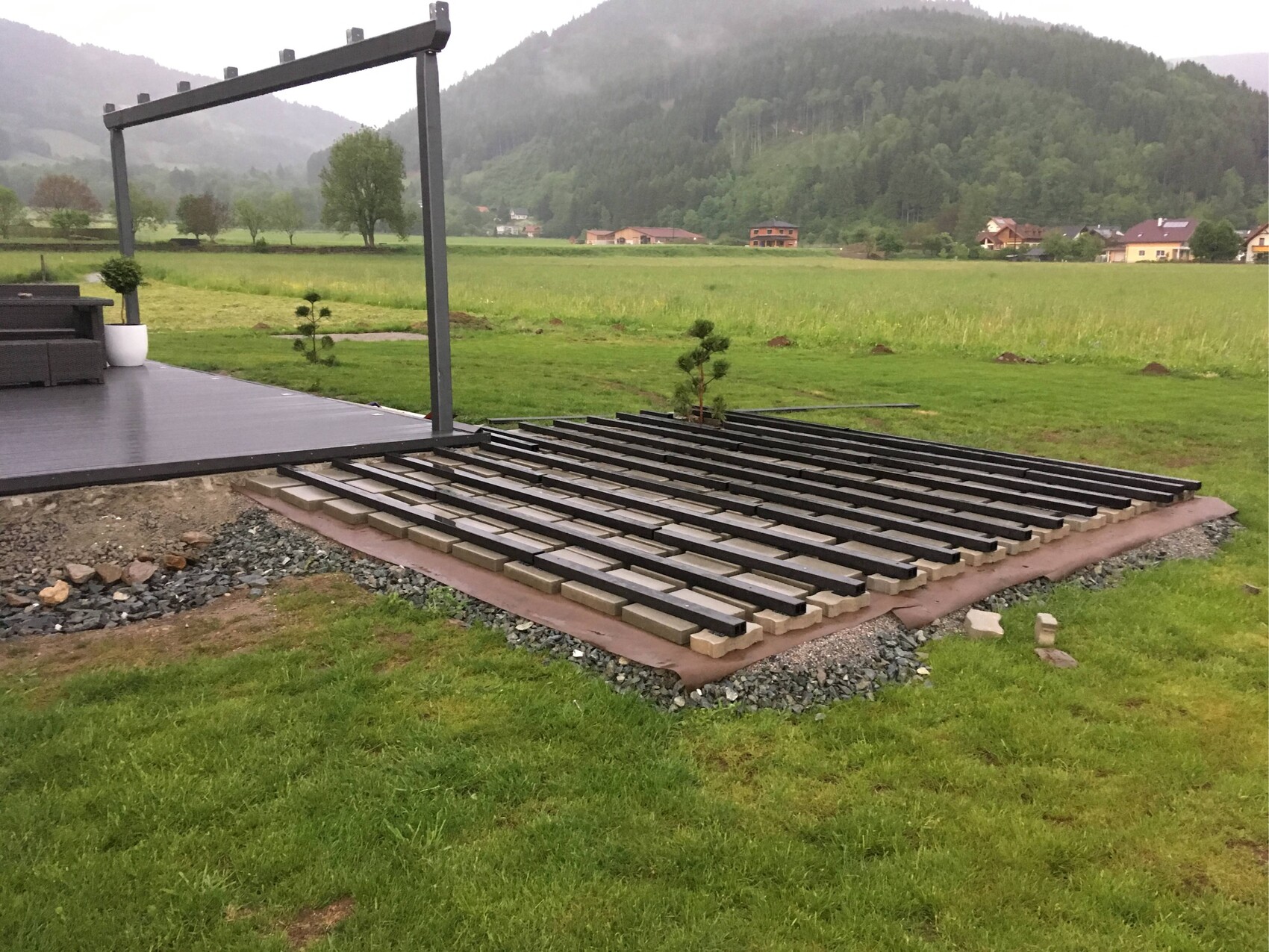 Moderne Outdoor-Terrasse mit Pergola, Holzlattenboden, grünem Feld und Berge im Hintergrund.