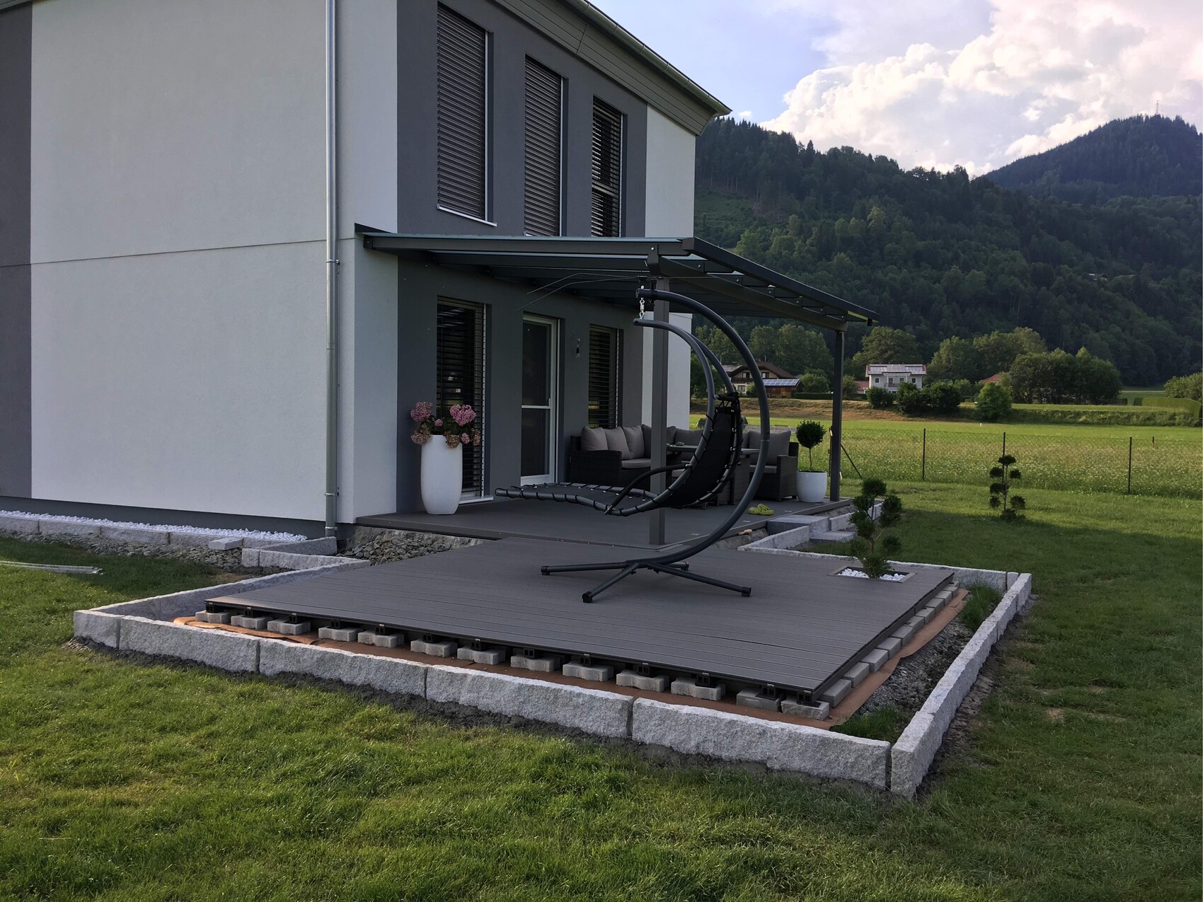 Moderne Haus-Terrasse mit grauer Lounge und Hängesessel; Wiese und Berge im Hintergrund.