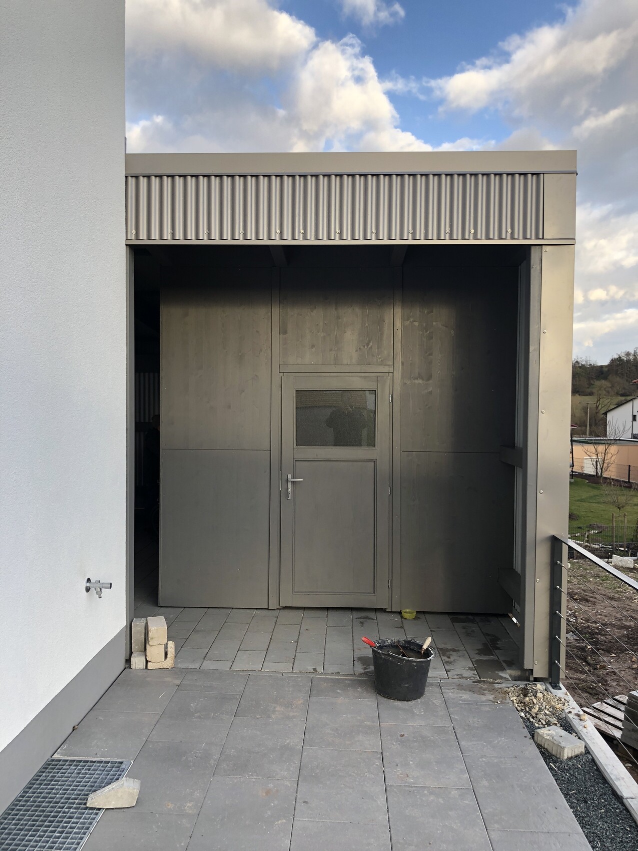 Grauer Metall-Vorbau mit Tür und Fenster auf Terrasse; Eimer, Ziegelsteine davor